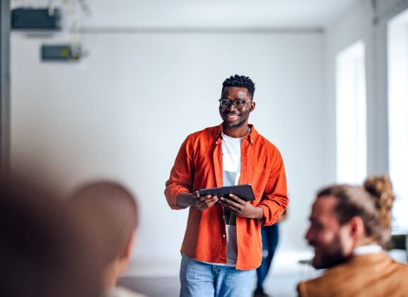 Handsome cheerful man in a orange shirt standing in front of an audience holding a tablet and using hand gestures to interact with the audience.