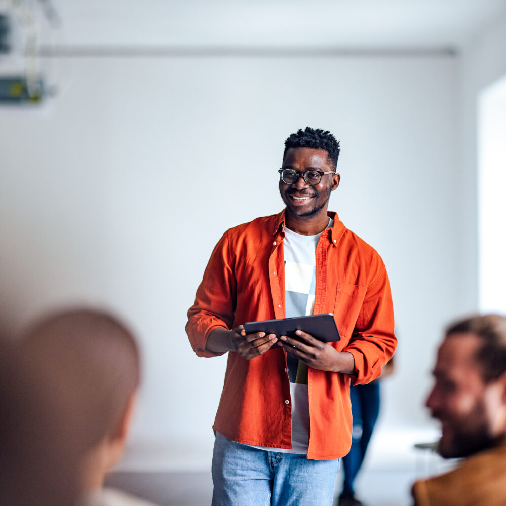 Handsome cheerful man in a orange shirt standing in front of an audience holding a tablet and using hand gestures to interact with the audience.