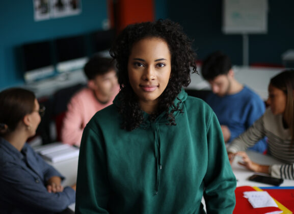 A young woman stands in focus in the foreground, smiling slightly at the camera, while a group of her peers sit in the background working together