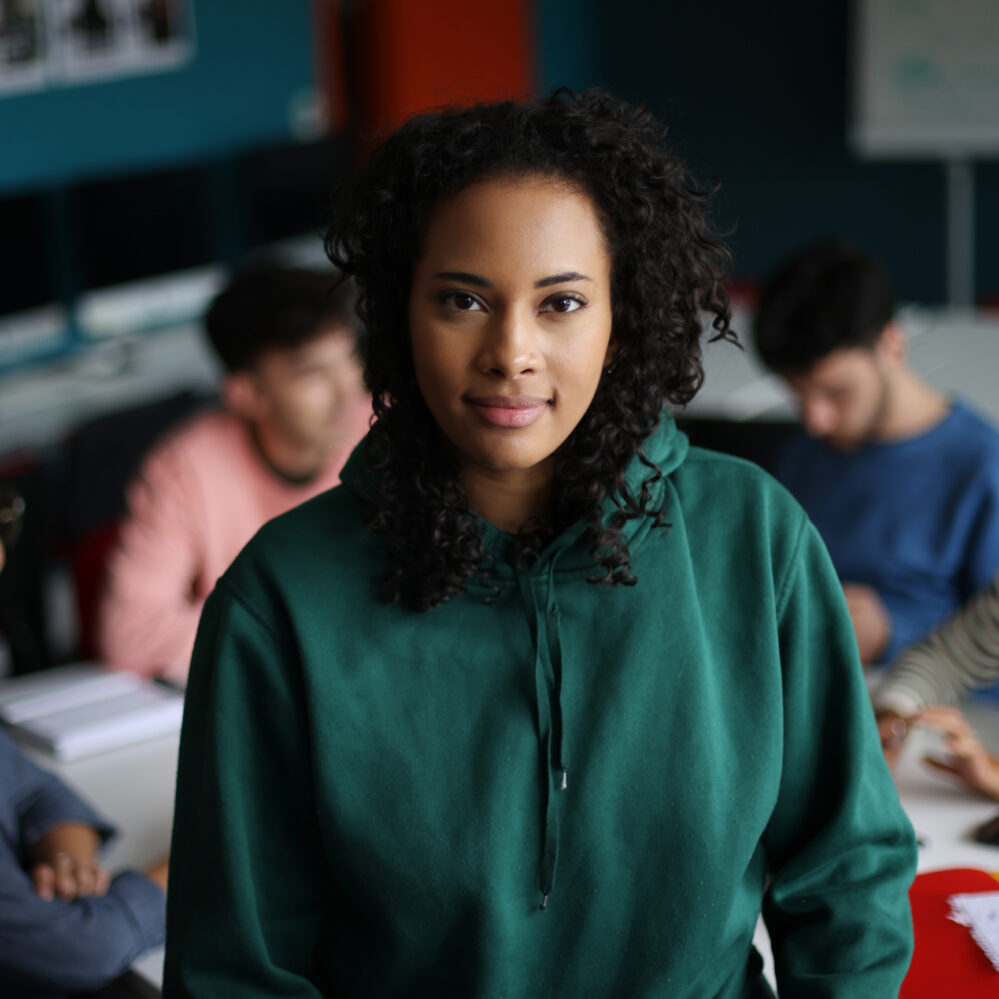 A young woman stands in focus in the foreground, smiling slightly at the camera, while a group of her peers sit in the background working together