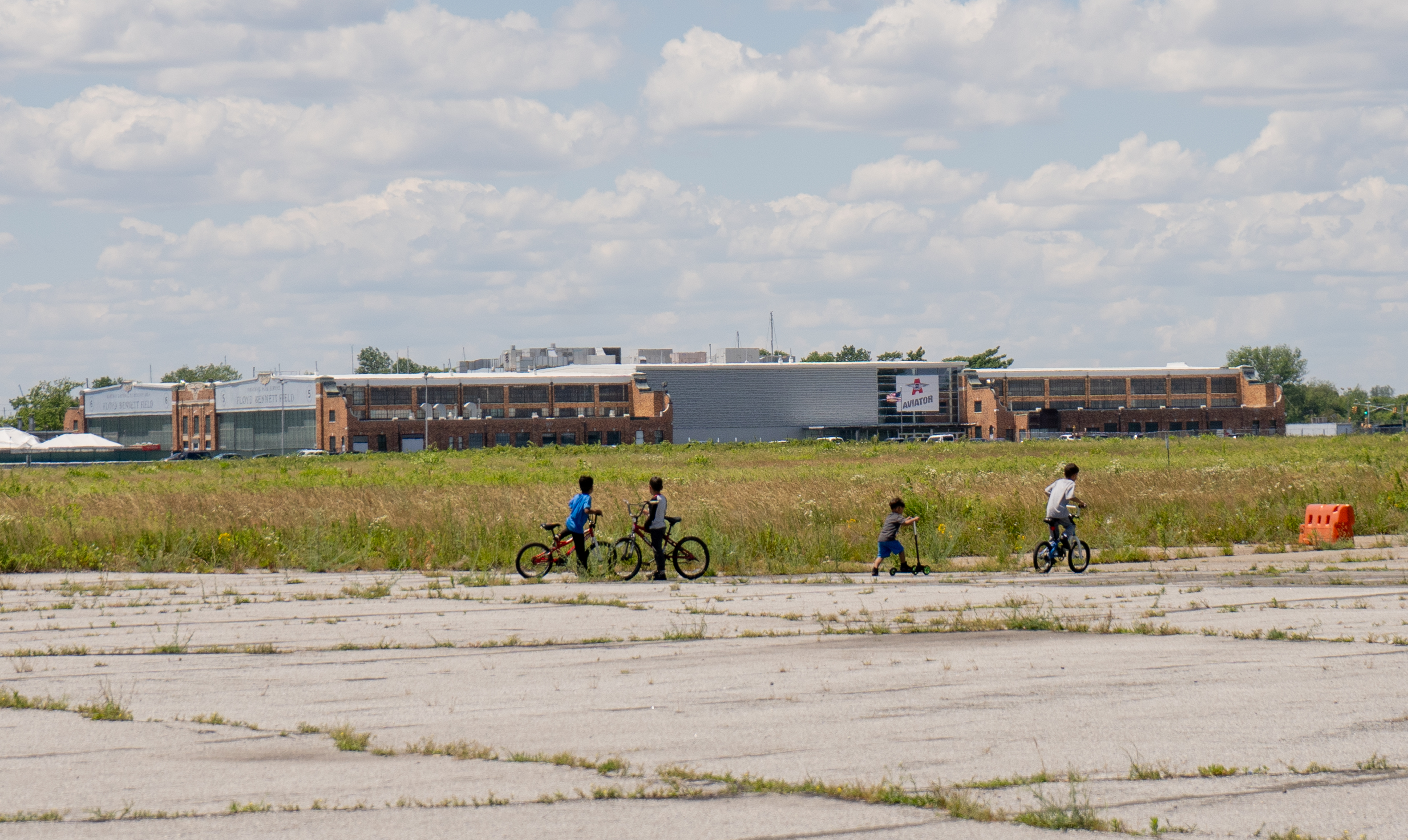 Image Caption: Young children of asylum seekers ride bicycles in an empty field outside their shelter at Floyd Bennett Field. Photo by Rudrani Ghosh.