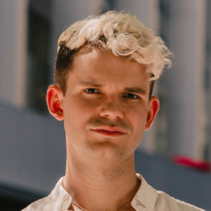 Portrait of Levi Bohanan. He has bleach blond curly hair and a beige collared shirt.