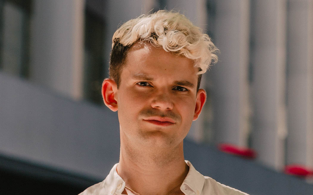 Portrait of Levi Bohanan. He has bleach blond curly hair and a beige collared shirt.