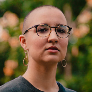 Portrait of Marcela Mulholland. She has a shaved head, tortoise shell glasses, and dangling gold earrings.