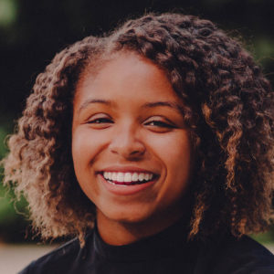 Portrait of Roquel Crutcher, she has a short curly hairstyle, a black shirt, and a wide smile.