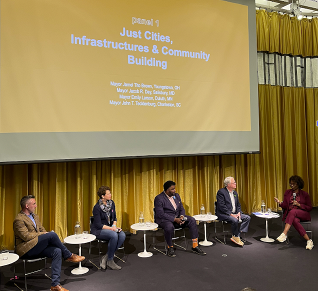 Panel 1 at The Mayors Imagining the Just City Symposium titled “Just Cities, Infrastructures and Community Building.” Five people sit in a row in an auditorium, directly in front of a large gold curtain and screen with the title of the panel projected onto it.